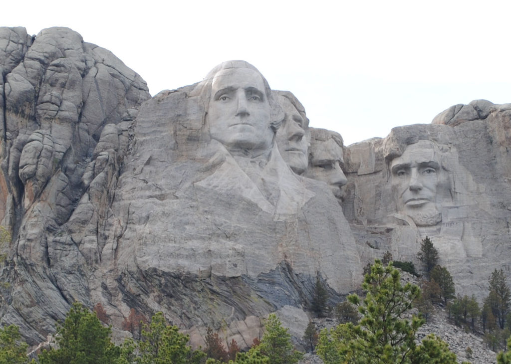 Mount Rushmore National Memorial with the carved faces of Washington, Jefferson, Roosevelt, and Lincoln set against rugged granite cliffs and pine trees under a clear sky.