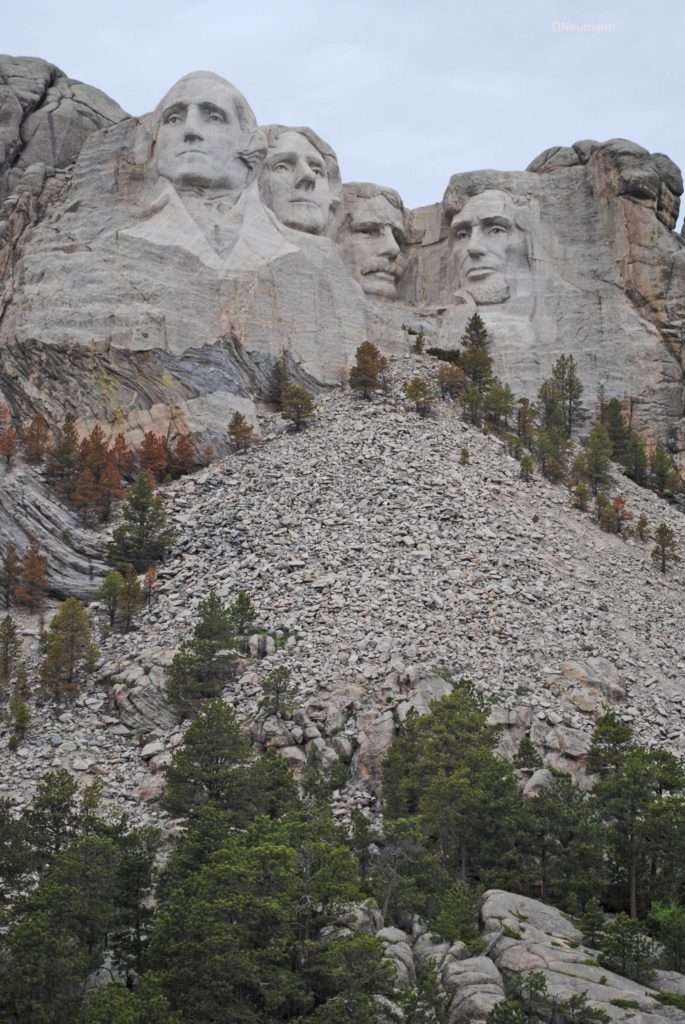 Wide view of Mount Rushmore showing the four presidential carvings above a large slope of loose shale and rock debris, with pine trees surrounding the base under an overcast sky.