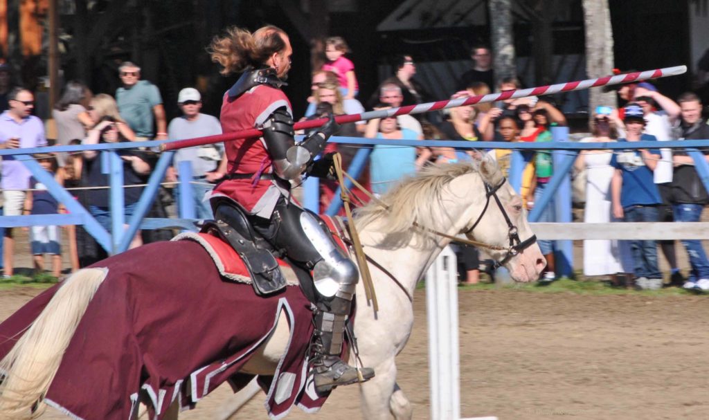 Armored jouster riding a white horse draped in maroon and white cloth, holding a red‑and‑white striped lance. The rider wears a red tunic over armor. Spectators stand behind a blue wooden fence watching the event at an outdoor renaissance fair.”