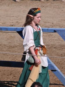“Person dressed in a green medieval-style outfit with a white undershirt, brown belt, and pouch, wearing pointed elf-like ears and a leafy headband. They are walking outdoors on a sandy surface near a blue wooden fence at a renaissance fair.”