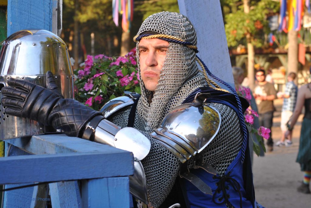 “Person dressed in medieval-style armor with chainmail and metal shoulder plates, resting a hand on a polished helmet placed on a blue wooden structure. Colorful flags and people appear in the background at a renaissance fair setting.”