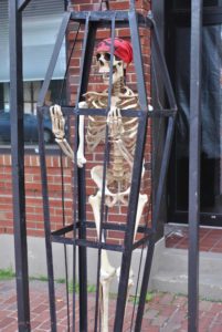 Skeleton figure wearing a red bandana posed inside a tall, narrow black metal cage outdoors. The cage sits on a brick walkway in front of a red‑brick building, with the skeleton gripping the bars as part of a Halloween or spooky decorative display.