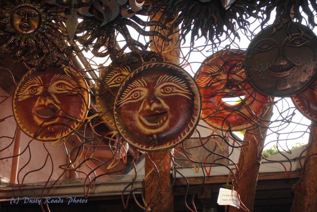 A display of decorative metal sun sculptures hanging together at a Key West market, each with a stylized face and radiating flame‑like rays in bronze, copper, and gold tones.