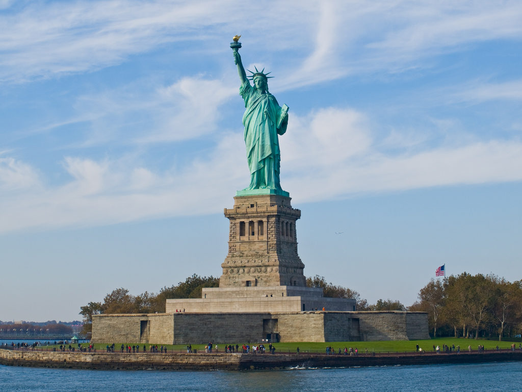  The Statue of Liberty standing on its pedestal on Liberty Island, with visitors below, an American flag to the right, and a partly cloudy sky overhead.
