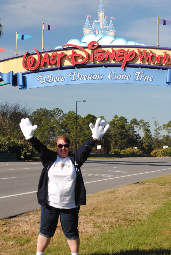 A woman wearing oversized white cartoon gloves, sunglasses, a white shirt, and a dark jacket stands on a grassy roadside with her arms raised in front of the Walt Disney World entrance sign that reads “Where Dreams Come True.” Colorful flags and a castle graphic top the sign, capturing a playful and celebratory moment at the park entrance.