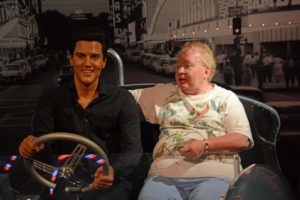 Visitor posing beside a wax figure seated in a vintage car display at Madame Tussauds, with a black‑and‑white city street backdrop.