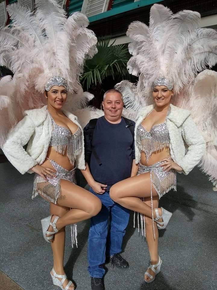 Tourist posing with two Las Vegas showgirls in silver sequined costumes and feathered headdresses, standing outdoors with palm trees in the background.