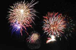 Colorful fireworks burst against a dark night sky, with red, blue, white, and gold streaks radiating outward in bright, celebratory patterns.