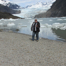 A person stands on a rocky shoreline facing floating ice and a massive glacier stretching between snow‑covered mountains under a cloudy sky at Mendenhall Glacier in Alaska.