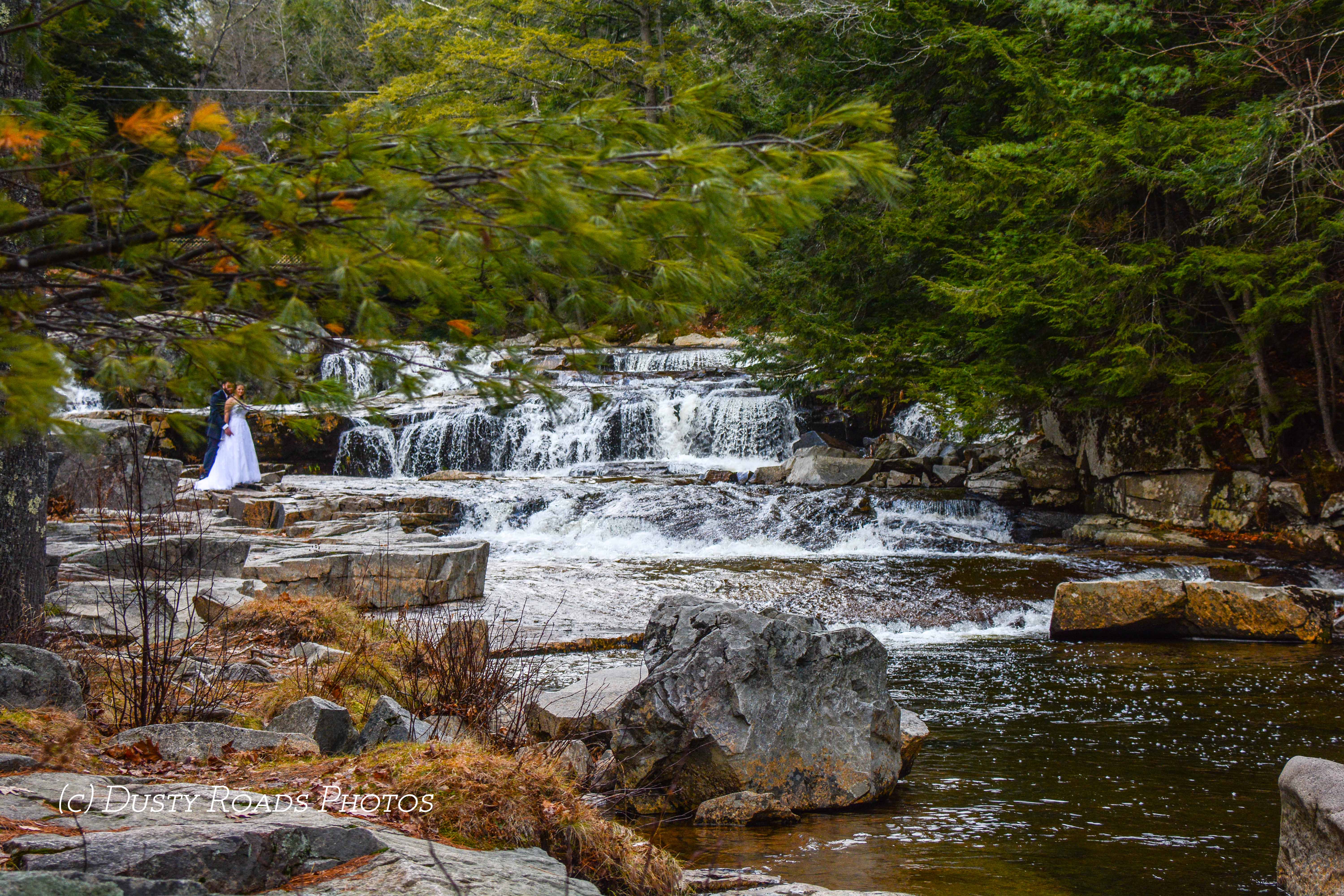 A bride in a white gown and a groom in a dark suit stand on rocks beside a small waterfall surrounded by evergreens, with autumn foliage and natural stone creating a peaceful New England outdoor wedding scene.