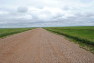 Straight dirt road stretching across wide open grassy plains under a cloudy sky, disappearing toward the distant horizon.
