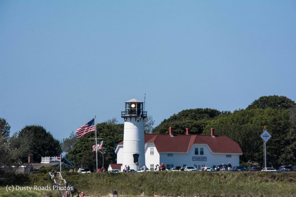 Chatham Light on a clear summer day, the white tower and red‑roofed Coast Guard station standing against blue sky and green trees, with visitors gathered out front.