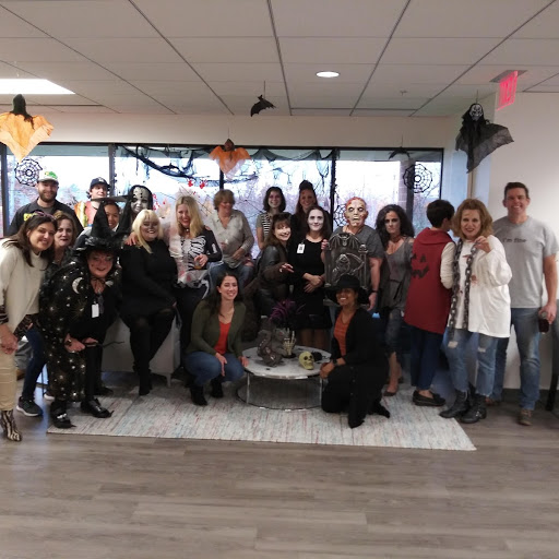 Group of people dressed in Halloween costumes—including witches, skeletons, and other spooky characters—posing indoors in a room decorated with bats, cobwebs, and orange ornaments. A small round table with Halloween props like a skull and a spider sits in the center, creating a festive party atmosphere.