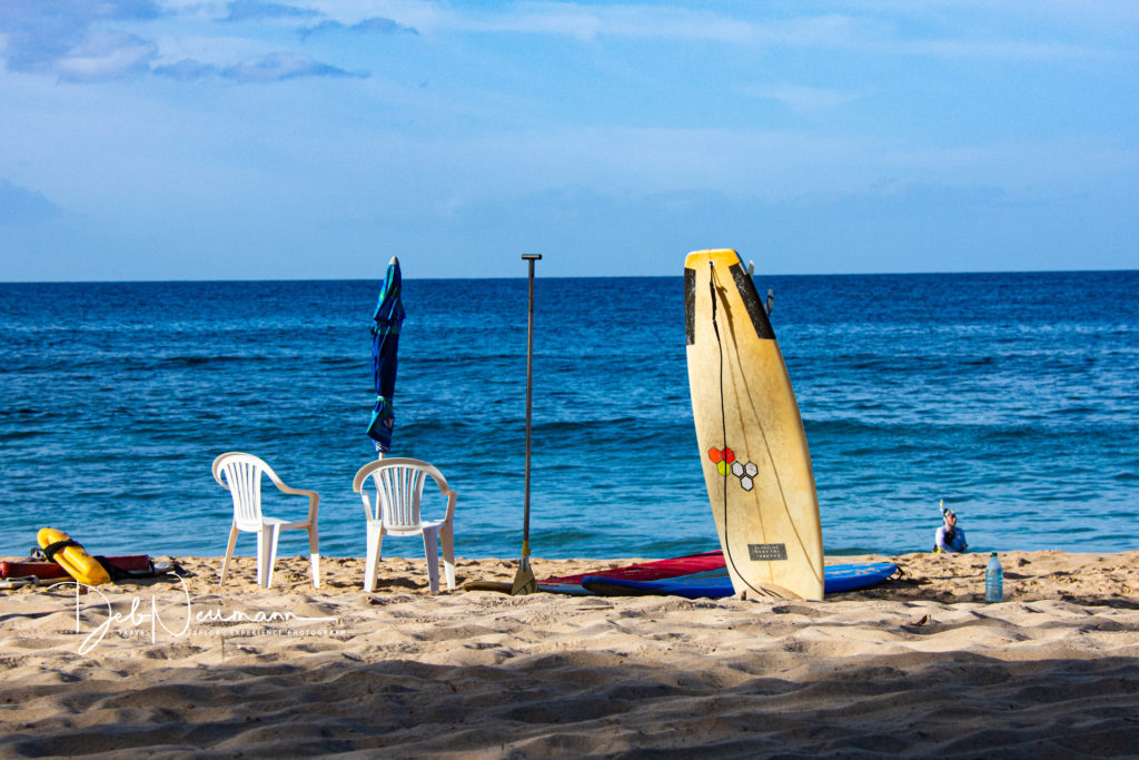 Two white beach chairs sit on sandy shore facing a calm ocean. A folded blue umbrella and paddle stand nearby. In the foreground, two surfboards rest in the sand, one upright with colorful hexagonal decals. A person sits near the water’s edge under a bright sky with soft clouds