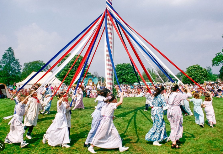 “Kids holding colorful ribbons as they dance around a tall maypole on a sunny May Day, with families watching from the crowd.”