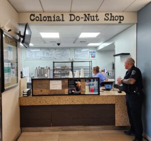 A police officer stands at the counter of the Colonial Do‑Nut Shop kiosk inside Morton Hospital, with staff preparing donuts behind the counter.