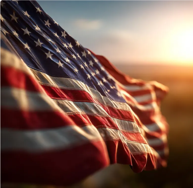 : An American flag waves in the wind against a warm golden sunset, its red and white stripes glowing in the light and the blue field of stars clearly visible.