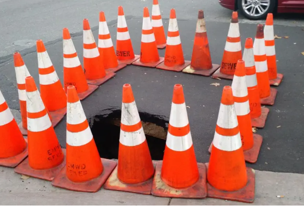 A tight cluster of bright orange traffic cones surrounding a fresh sinkhole in a Boston street, with a red car parked nearby.