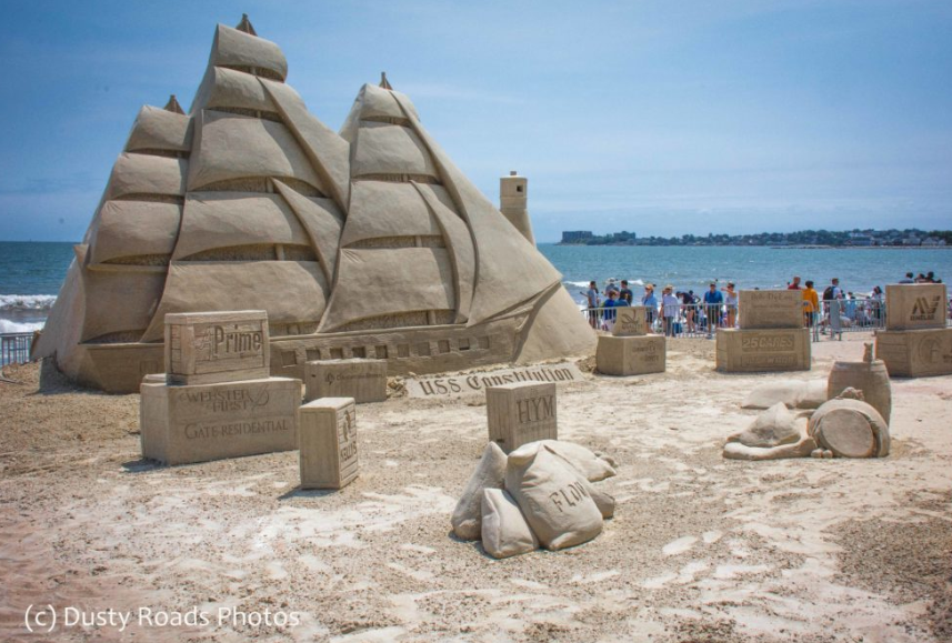 A detailed sand sculpture of the U.S.S. Constitution on Revere Beach, surrounded by carved crates and barrels, with beachgoers and the ocean in the background.