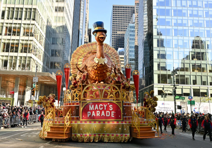The giant turkey float wearing a blue top hat in the Macy’s Thanksgiving Day Parade, moving past crowds and tall city buildings.”