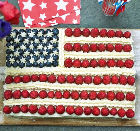 A rectangular 4th of July cake decorated like the American flag, with blueberries and small white star-shaped frosting in the top-left corner and alternating rows of raspberries and white frosting forming the stripes. The cake sits on a wooden surface with patriotic napkins and red, white, and blue striped straws in the background.