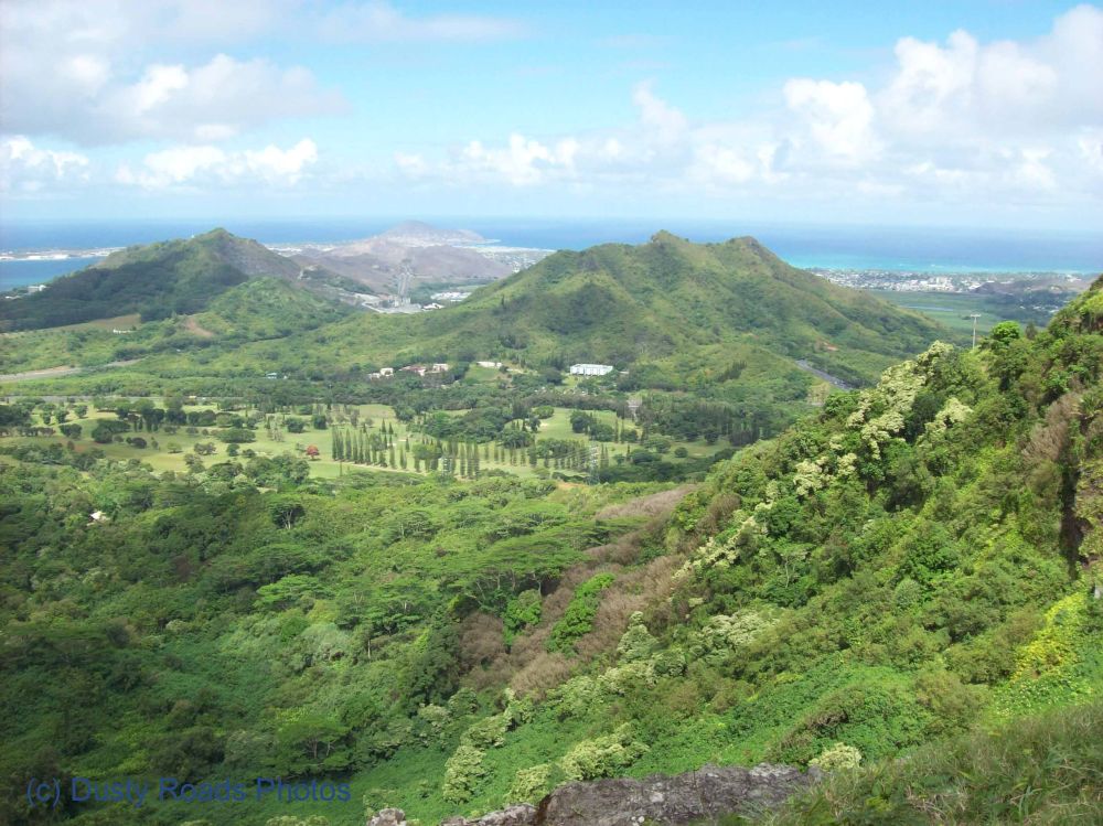 From the windy heights of Nuʻuanu Pali, the view opens to a deep green valley and the rugged Koʻolau Mountains, with Honolulu and the shimmering coastline far beyond.