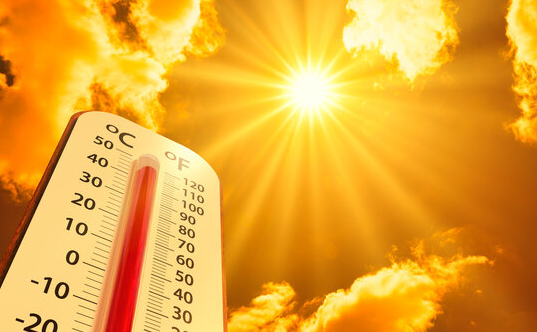 A thermometer showing both Fahrenheit and Celsius scales rises close to the top against a bright, golden sky. Sunlight flares behind scattered clouds, emphasizing extreme heat and a hot summer day.