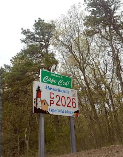 A roadside “Welcome to Cape Cod” sign surrounded by tall green trees, featuring a lighthouse illustration and the text “Massachusetts – Cape Cod & Islands – 2026.”