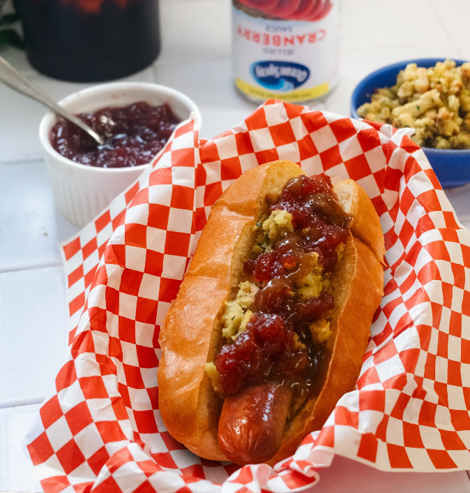 A hot dog in a bun topped with stuffing and cranberry sauce, served in a red‑and‑white checkered basket with bowls of stuffing and cranberry sauce in the background.