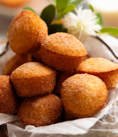 Mini cinnamon‑sugar donut muffins nestled in a parchment‑lined bowl, their tops glistening with sugar, with soft greenery and a white flower behind them.