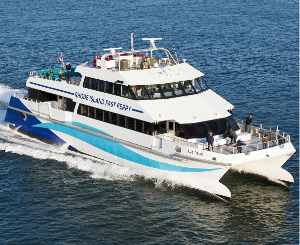 A high‑speed passenger ferry named “Ava Pearl” cuts across the water on a sunny day, carrying travelers on its open upper deck. The white and blue catamaran creates a wide wake behind it as it heads toward Martha’s Vineyard.