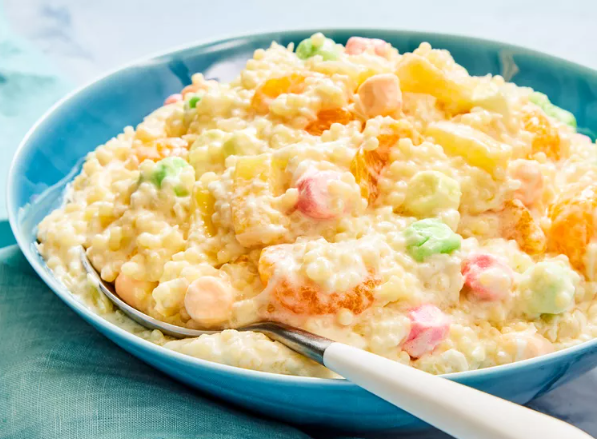 A blue bowl filled with creamy frog eye salad made of acini di pepe pasta, pineapple, mandarin oranges, and colorful mini marshmallows, with a spoon resting inside the mixture.