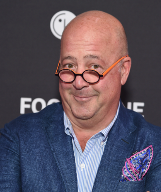Andrew Zimmern posing at a media event, wearing round orange glasses, a blue blazer, a striped shirt, and a colorful pocket square against a dark backdrop with partial text.