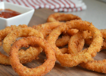 Golden, crispy onion rings piled on a wooden surface with a small dish of red dipping sauce in the background.”