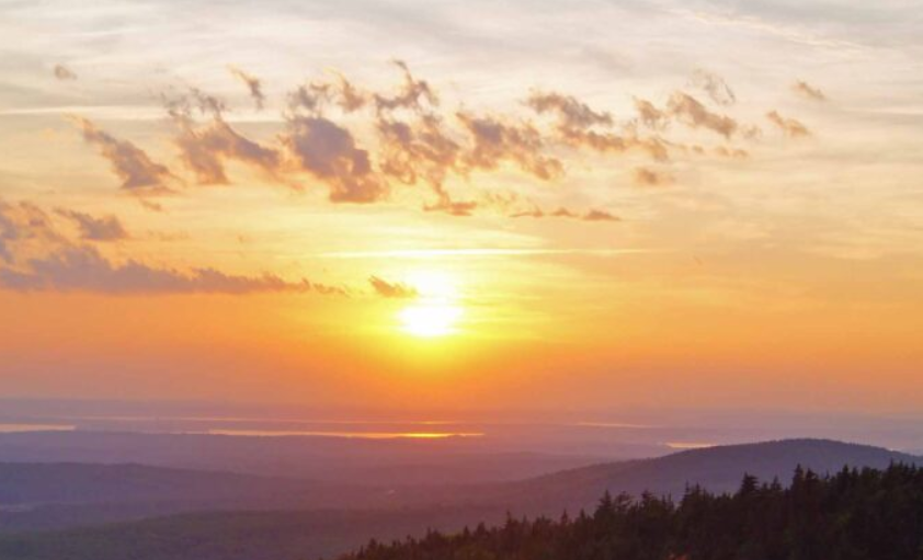 Sunset over Cadillac Mountain in Maine, with warm orange light spreading across layered hills and a forested foreground beneath a glowing sky.
