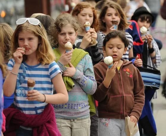 Children walking outdoors in cool weather, happily eating ice‑cream cones, with buildings blurred in the background.