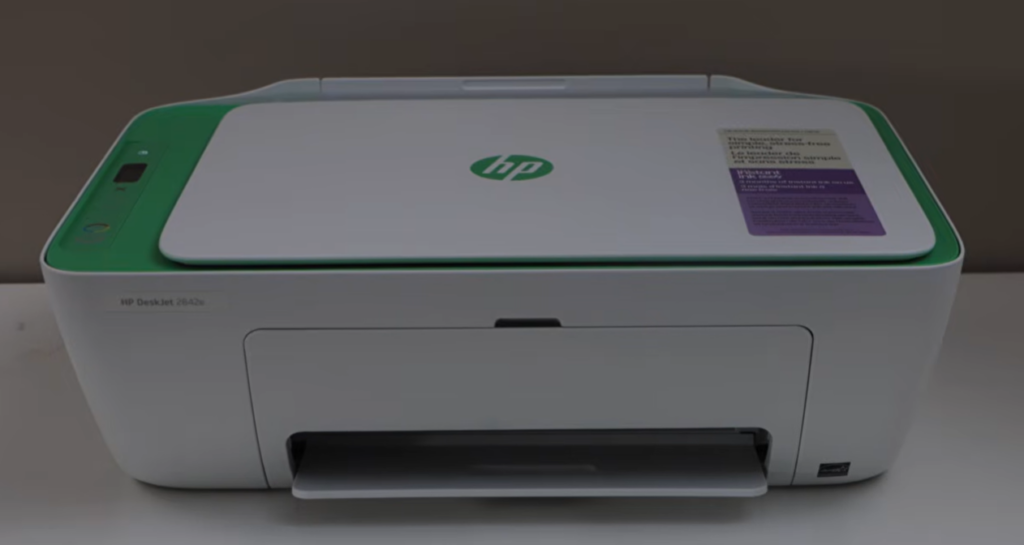 A white HP DeskJet 2842e printer with green accents sits on a light surface. The top panel shows the HP logo, control buttons, and a setup sticker. The printer looks compact and slightly mischievous — the perfect visual for “Mr. Cranky Pants,” the stubborn printer from my technology saga.