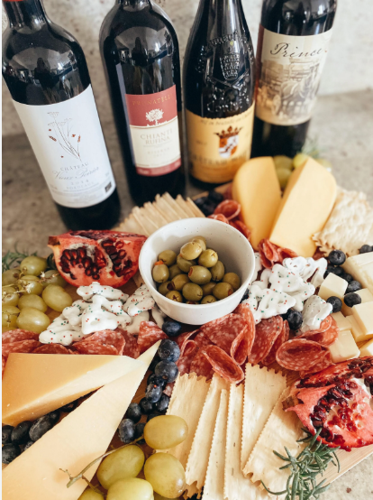 A charcuterie board arranged with assorted cheeses, sliced cured meats, green grapes, blueberries, pomegranate halves, crackers, green olives in a small bowl, and a creamy white dip. Behind the board are four bottles of red wine, creating an elegant wine‑and‑cheese presentation.