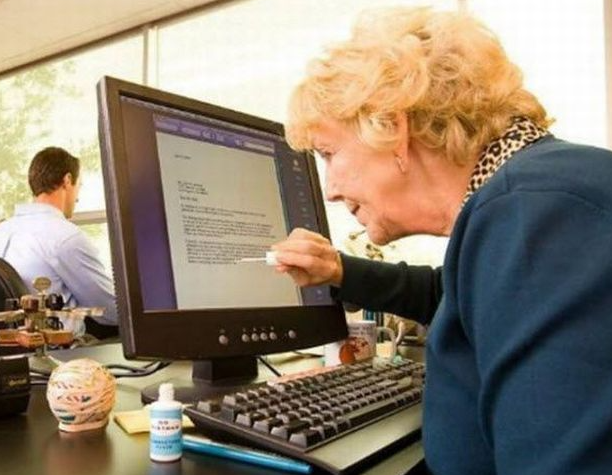 An older person sits at a desk in front of a computer monitor, holding a bottle of correction fluid and applying it directly onto the screen as if trying to fix a typo. The monitor displays a document with typed text. A keyboard, mouse, rubber band ball, and office supplies sit on the desk. In the background, another person works at a separate desk. The scene is humorous, showing a mix‑up between old‑school office tools and modern technology.