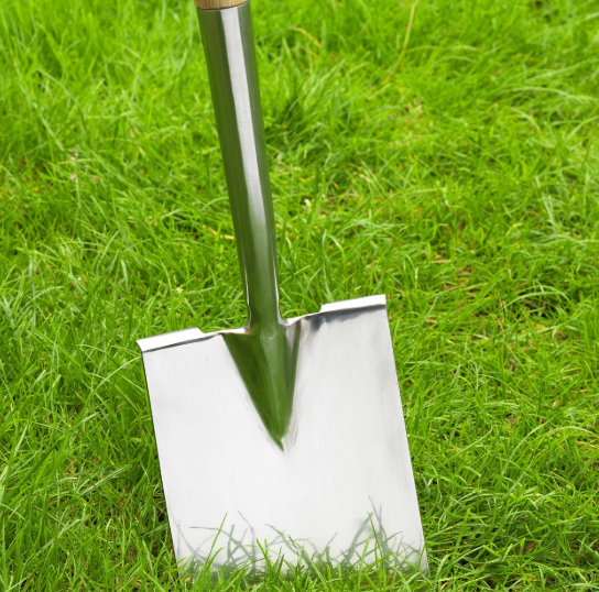 “A shiny metal spade with a wooden handle standing upright on bright green grass, its flat blade reflecting light.”