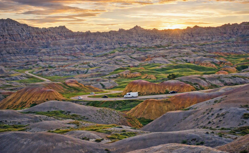Enhanced view of the Badlands showing colorful layered rock formations, rugged hills, and a winding road with cars and an RV traveling through the landscape under a partly cloudy sky.
