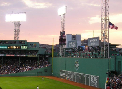 A sunset view of Fenway Park with the Green Monster in left field, packed stands, and an outfielder positioned near the wall under warm evening light.