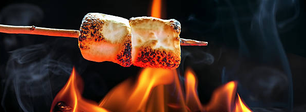 Two marshmallows on a wooden stick roast over bright orange flames, with the marshmallows turning golden brown as smoke rises from the fire.