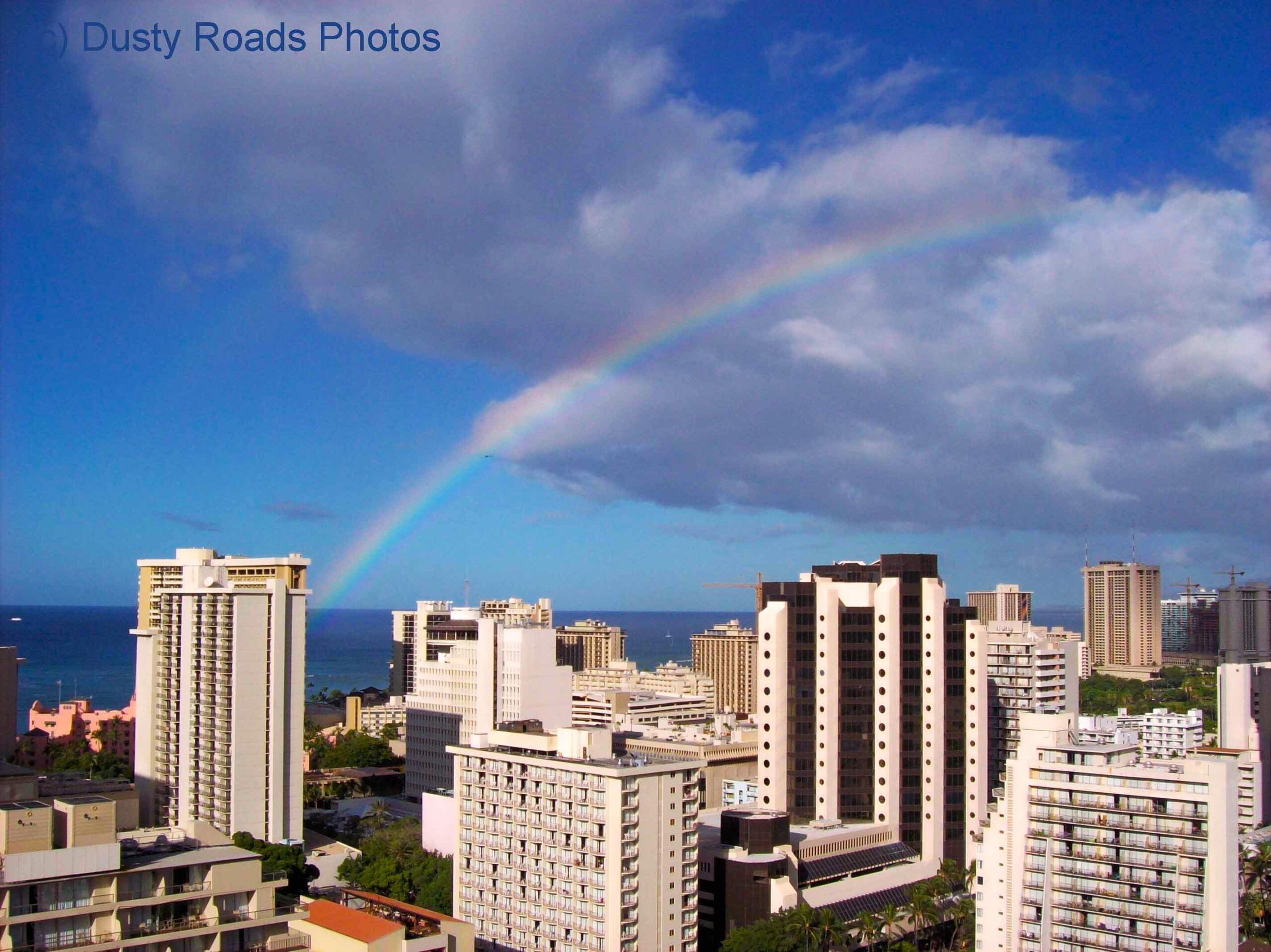 A rainbow arcs across the sky above Honolulu’s high‑rise skyline, with the ocean visible in the background. Sunlight breaks through scattered clouds, illuminating the tall buildings and creating a bright, colorful contrast against the cityscape.