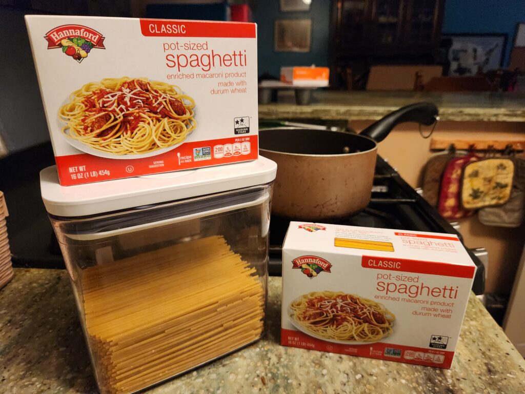 Two boxes of pot‑sized spaghetti on a kitchen counter beside a container of uncooked pasta, with a cooking pot in the background.