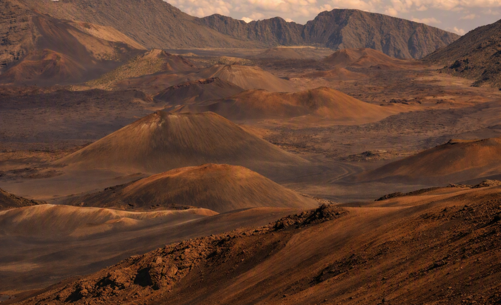 View from the summit of Haleakalā volcano on Maui showing reddish‑brown cinder cones and rugged volcanic terrain under a clear blue sky, with drifting clouds nestled among the crater slopes.”
