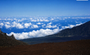   
“View from the summit of Haleakalā volcano on Maui showing rugged volcanic slopes and dark rocky terrain above a vast layer of white clouds under a deep blue sky.”