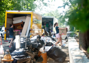 People loading furniture, boxes, and trash bags into large moving trucks on a Boston street during fall move‑in day.