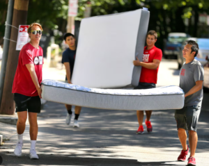  Four students carrying two mattresses down a sunny Boston street during college move‑in day.