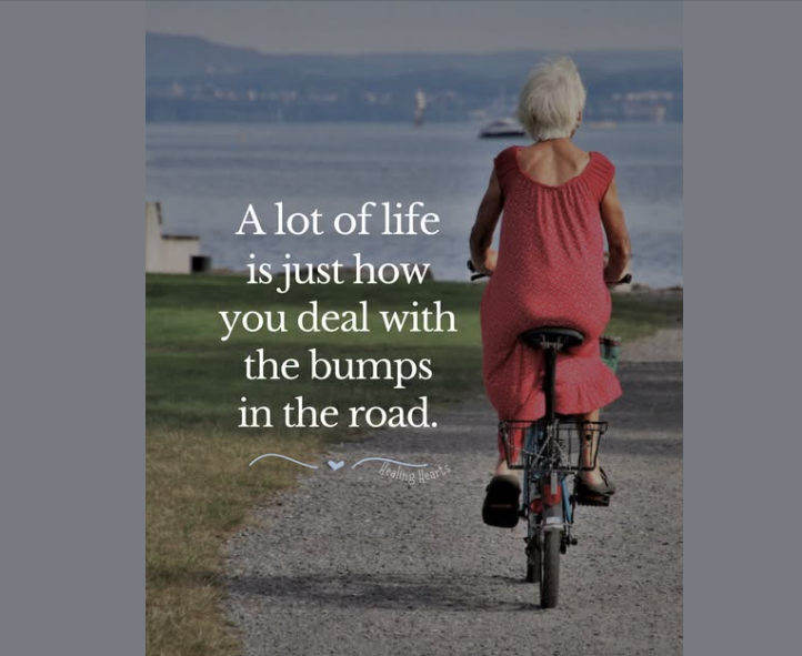 Older woman with short white hair riding a bicycle on a gravel path beside a lake, wearing a loose red dress, with the quote “A lot of life is just how you deal with the bumps in the road” and a small “Healing Heart’s” logo beneath it.